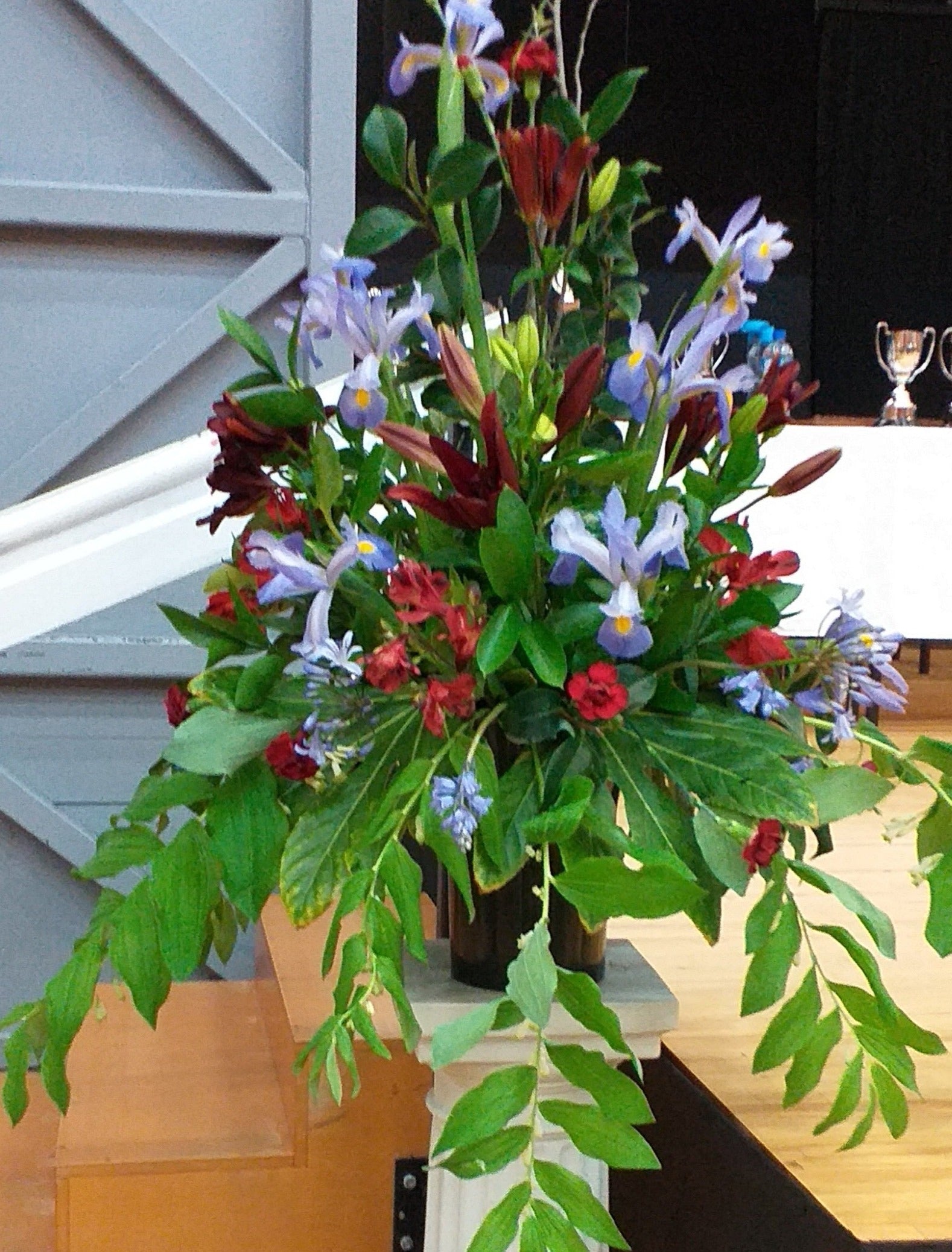 a pedestal arrangement in red and pale blue, including spray carnations, red lilies, red alstro and blue irises,