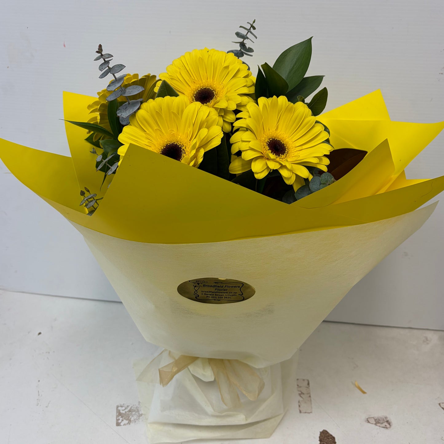 A bouquet of yellow gerbera daisies with green leaves and silver eucalyptus stems, wrapped in bright yellow paper and cream-colored vylene