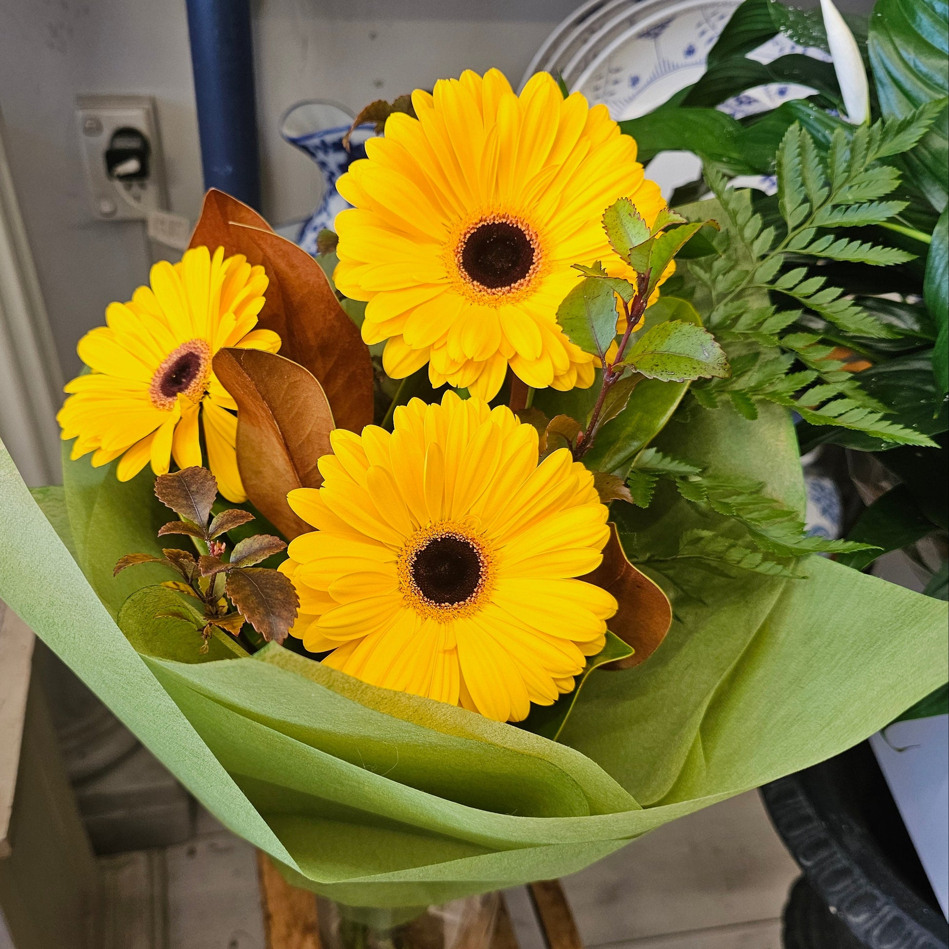 A bouquet of three bright yellow gerbera daisies with dark brown centers, wrapped in light green tissue paper  in a small glass vase 