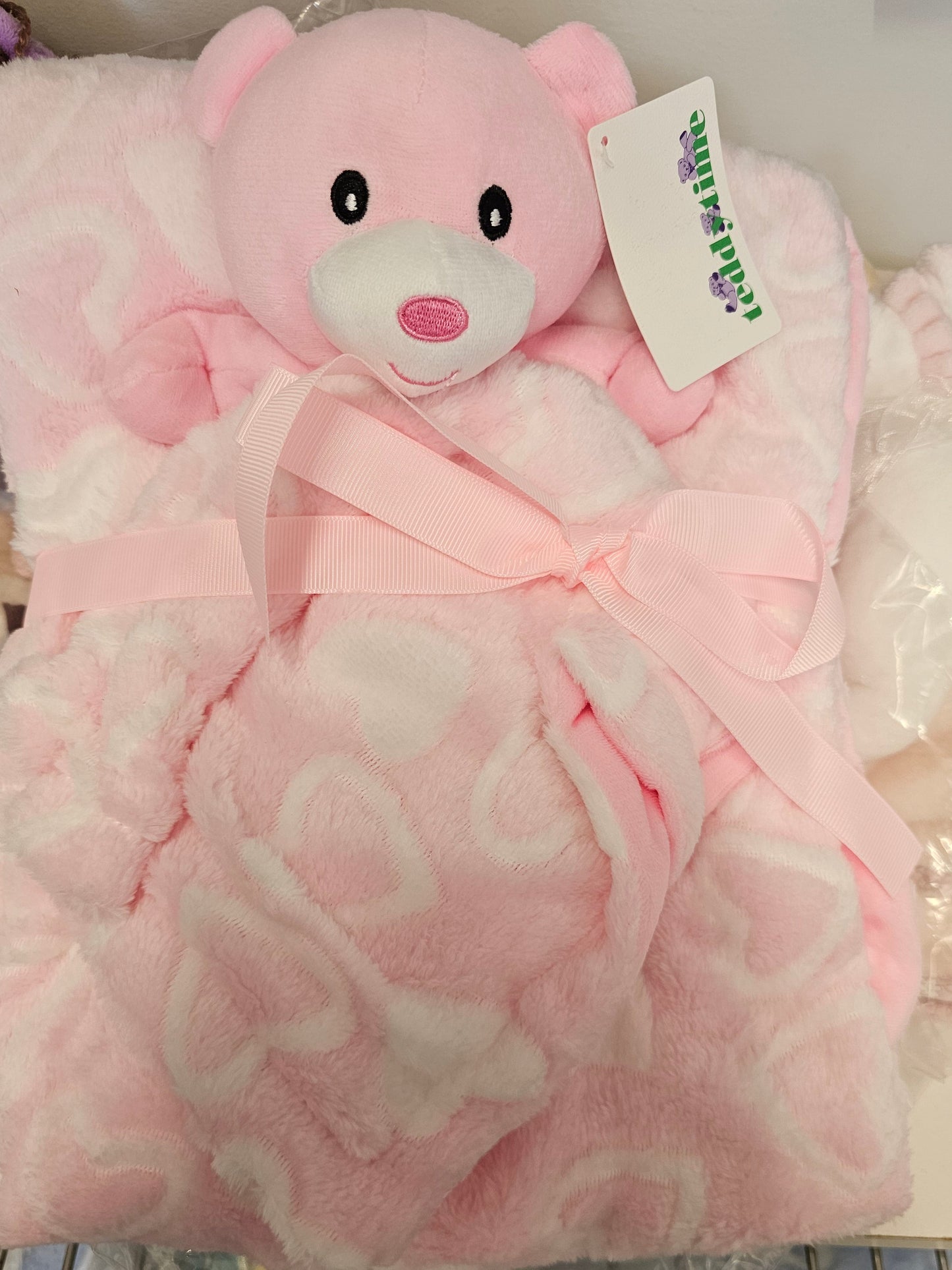 A pink plush baby blanket with a teddy bear face and a matching pink ribbon bow,
