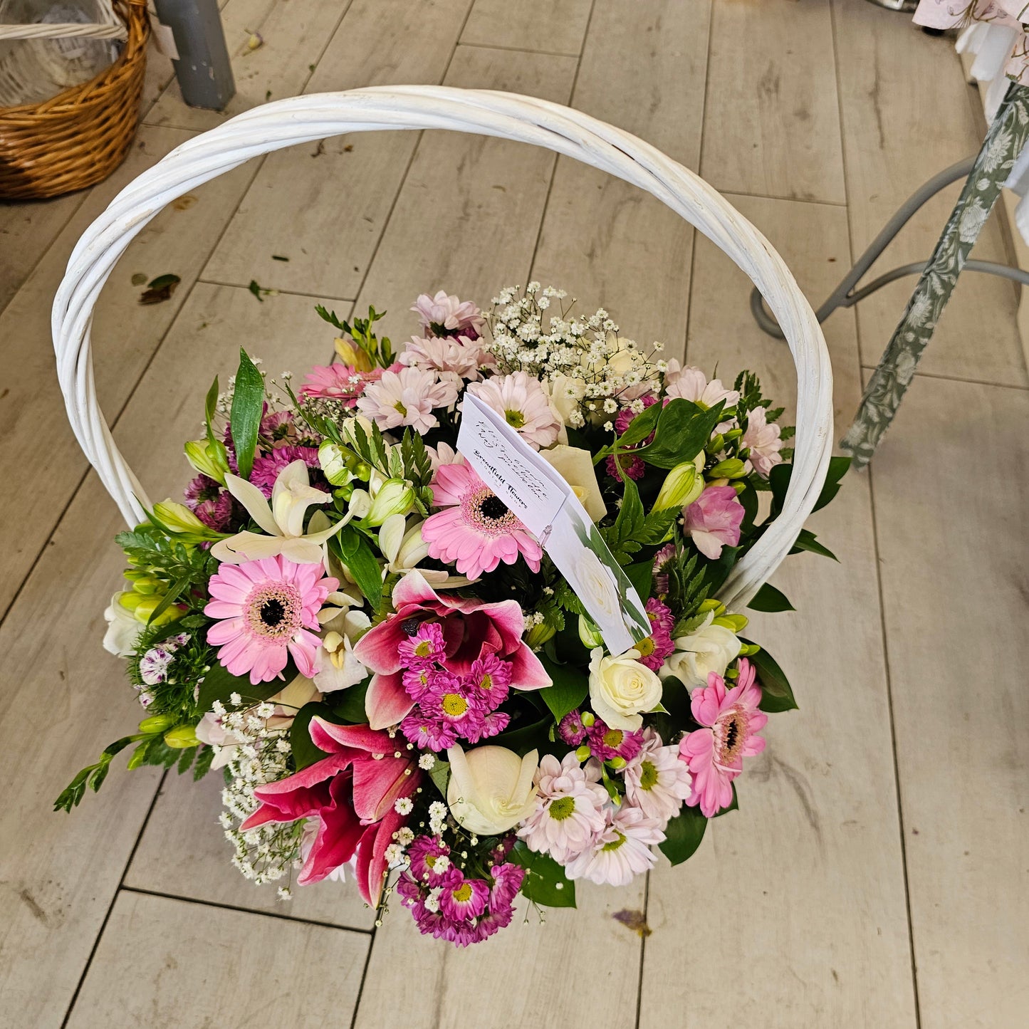 A round floral arrangement of pink, white, and green flowers in a white circular basket, photographed from above on a light wood floor.