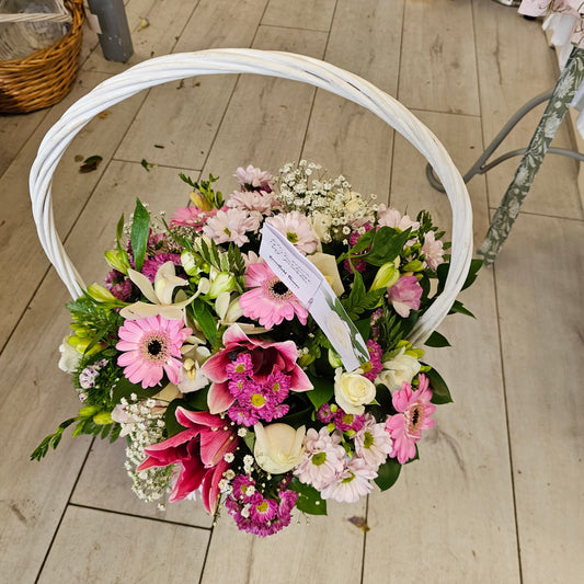 A round floral arrangement of pink, white, and green flowers in a white circular basket, photographed from above on a light wood floor.