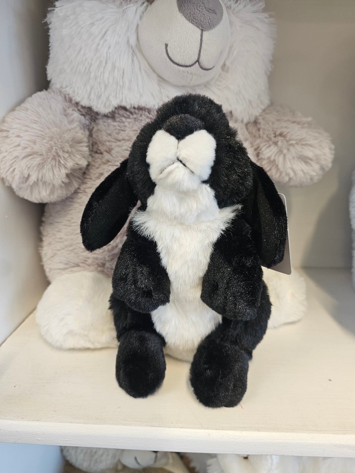 A black and white plush bunny toy lying on top of a light gray stuffed bear inside a white shelf or box.
