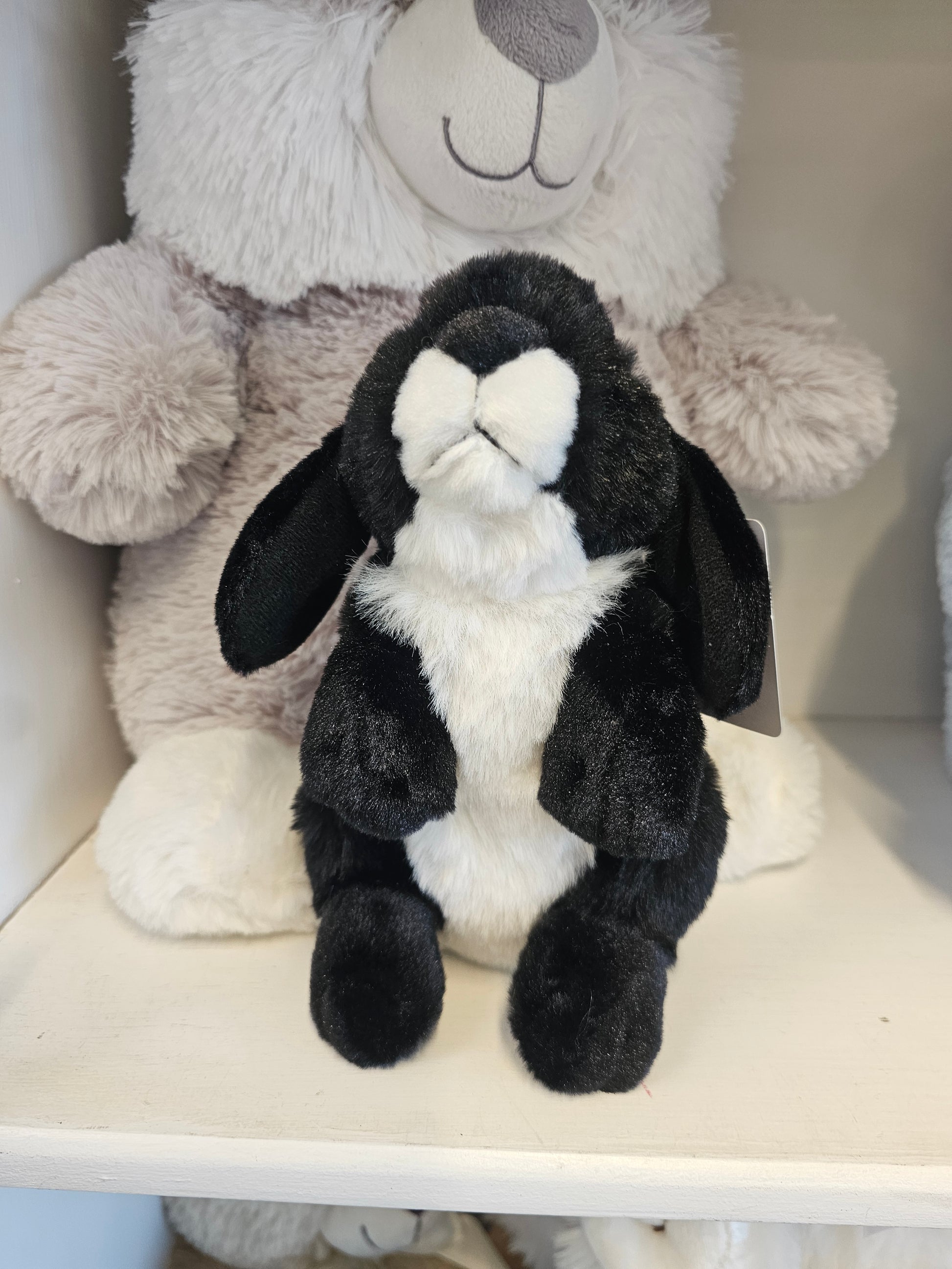 A black and white plush bunny toy lying on top of a light gray stuffed bear inside a white shelf or box.