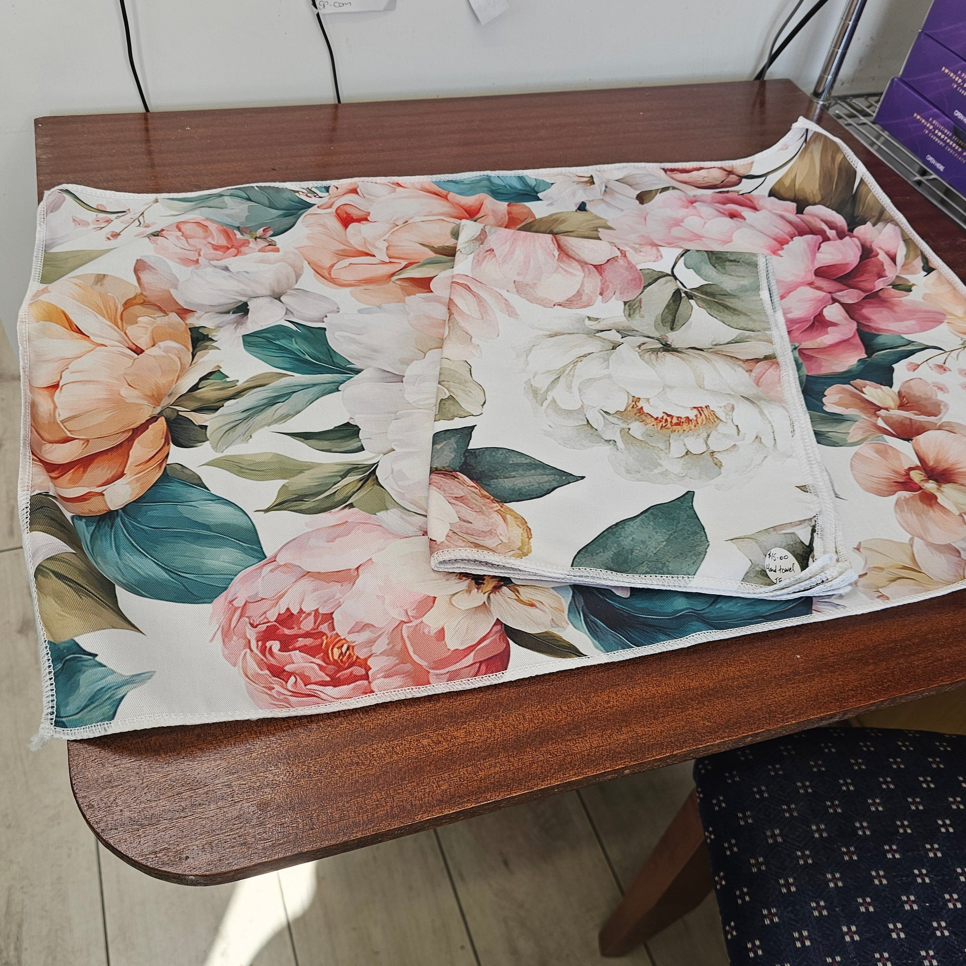 A floral-patterned fabric item, kitchen hand towel. The fabric features large watercolor-style peony flowers in pink, peach, and white with green leaves on a cream background.
