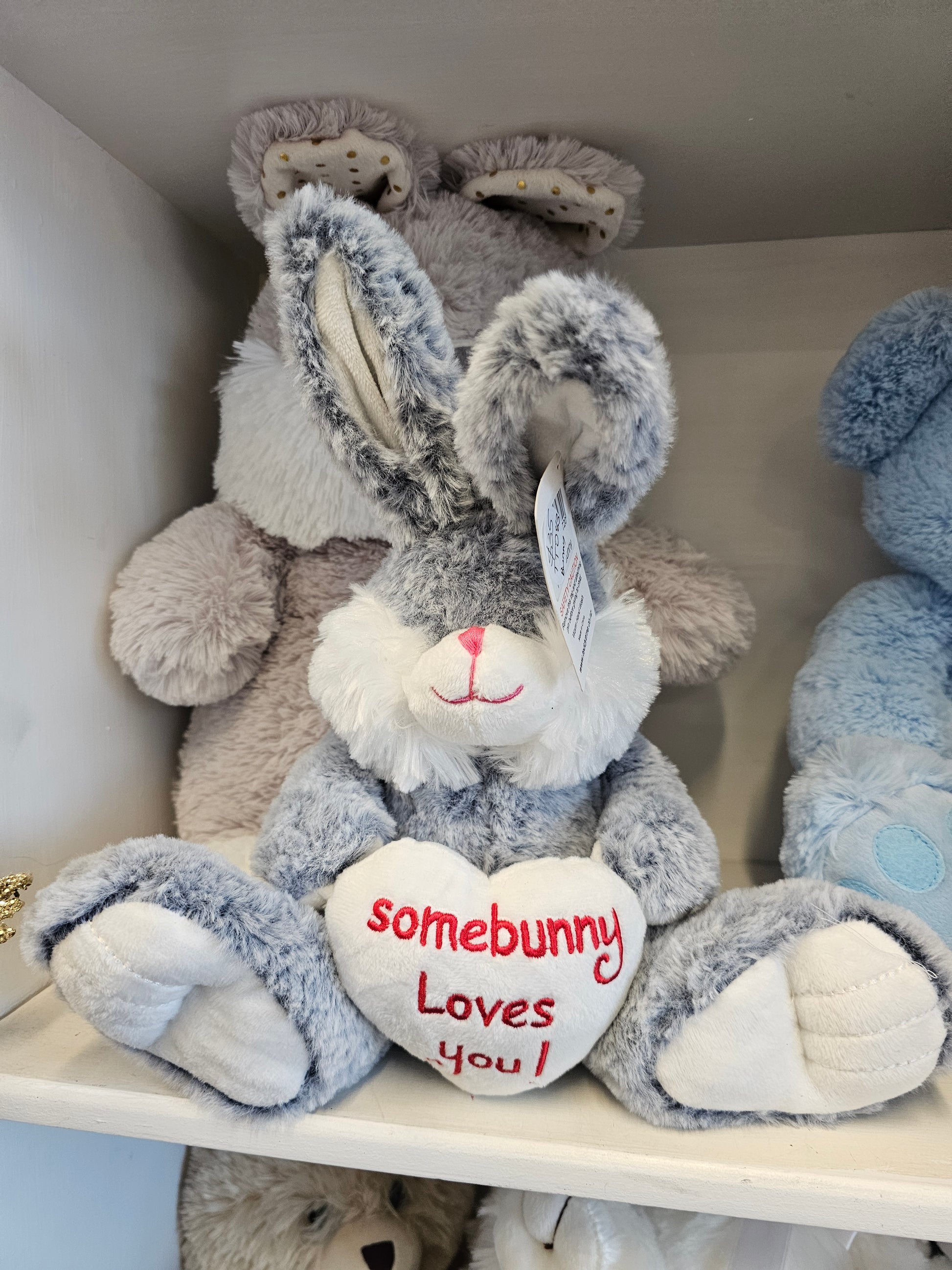 A plush gray and white stuffed bunny toy lying on a shelf, with a white heart patch that reads 'somebunny Loves you!' in red text.