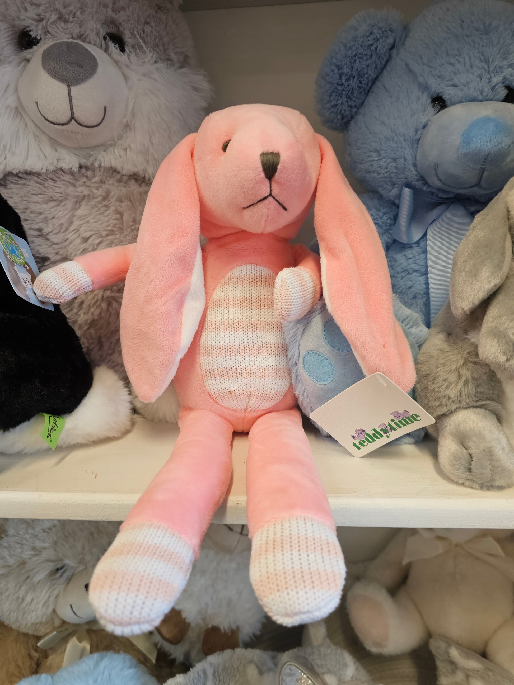 A pink plush bunny toy with long floppy ears, a striped belly, and white-and-pink striped socks, shown among other stuffed animals in a shelf display.