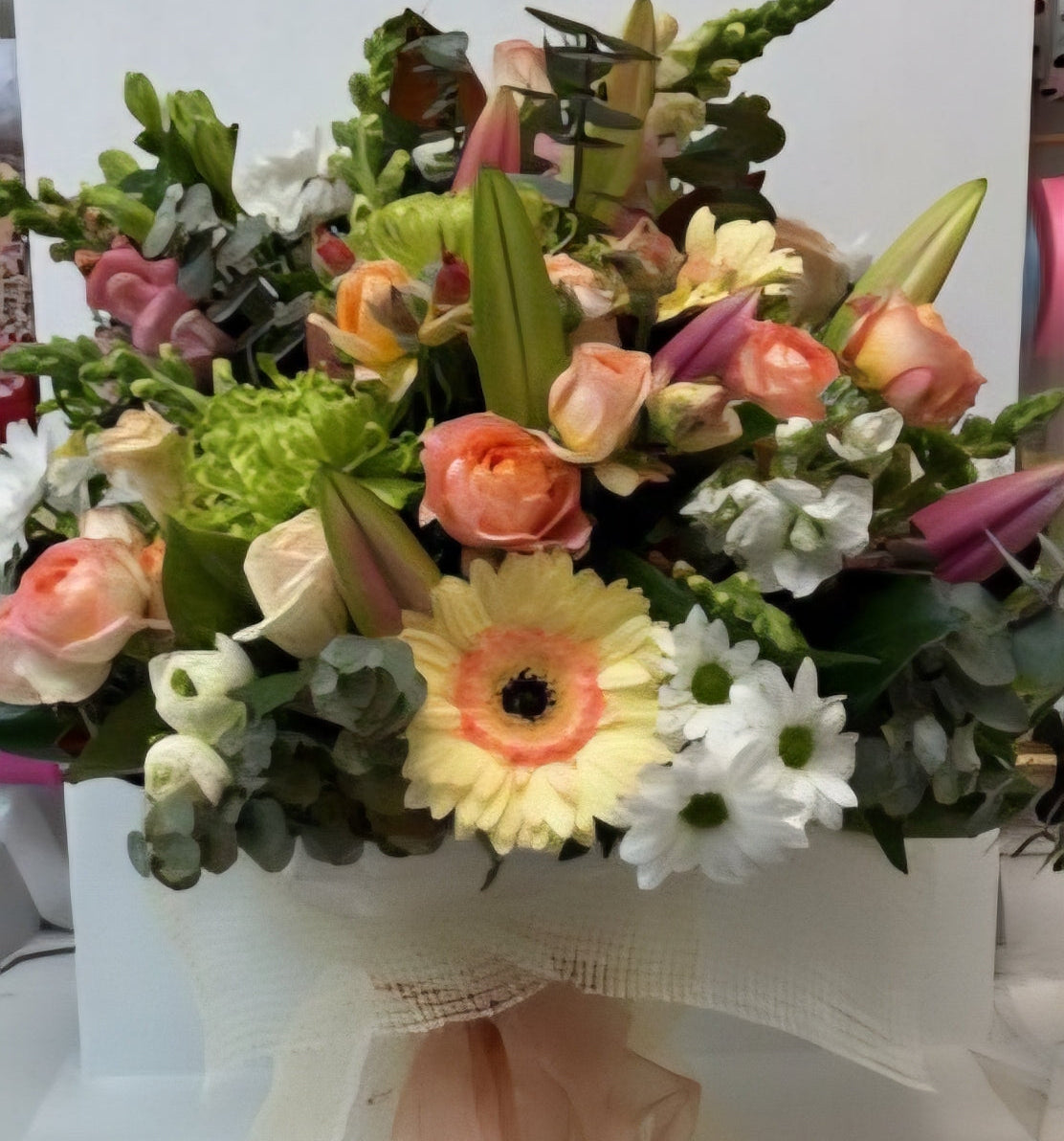 A vibrant mixed floral arrangement in a white box, featuring peach roses, a yellow and orange gerbera, white chrysanthemums, antirrhinums,pink lilies green chrysanthemums and green foliage, wrapped with a sheer peach ribbon.
