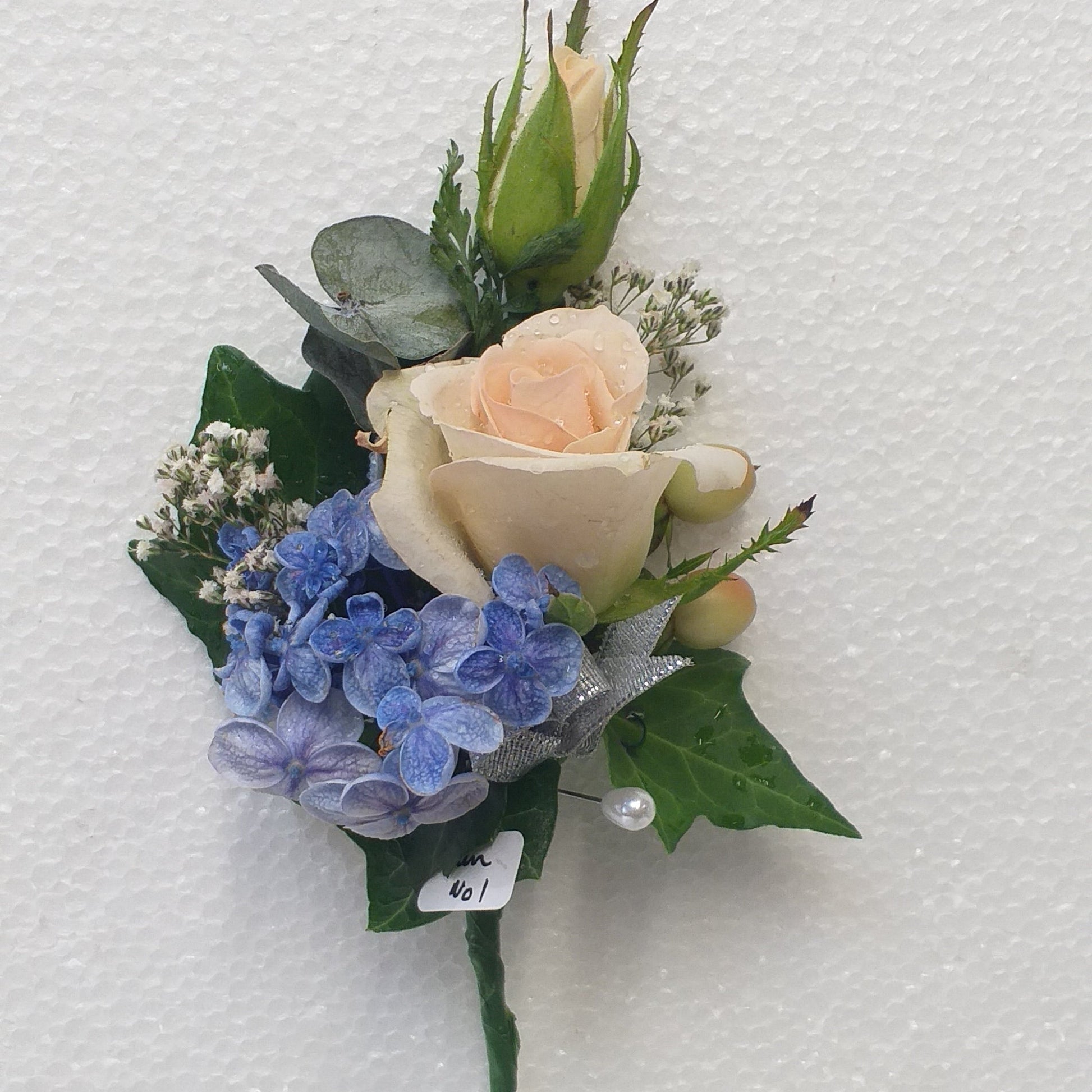 A small floral boutonniere featuring a pale peach rose, blue hydrangeas, green leaves, and a green rose bud