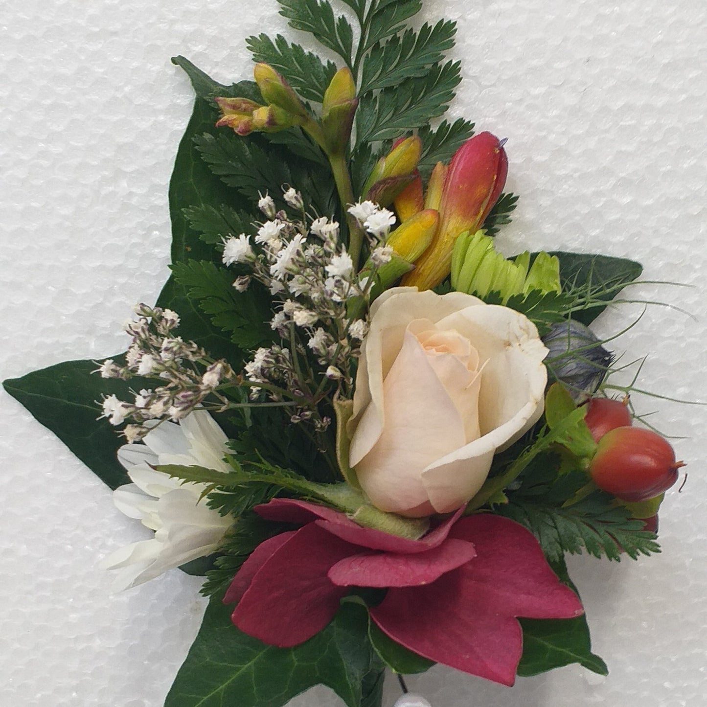 A floral wristlet or boutonniere arrangement featuring a pale peach rose, burgundy petal-like flowers, green leaves, small white baby's breath, and freesia buds