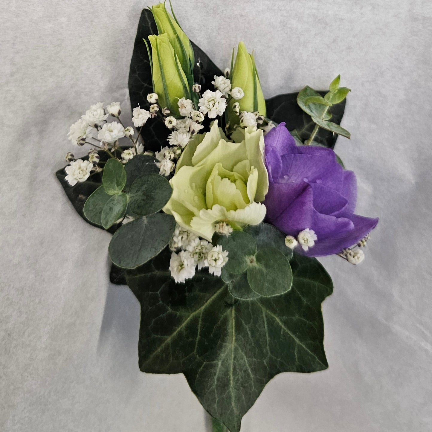 A small floral boutonniere with green ivy leaves, a pale green spray rose, a purple freesia, and white baby's breath