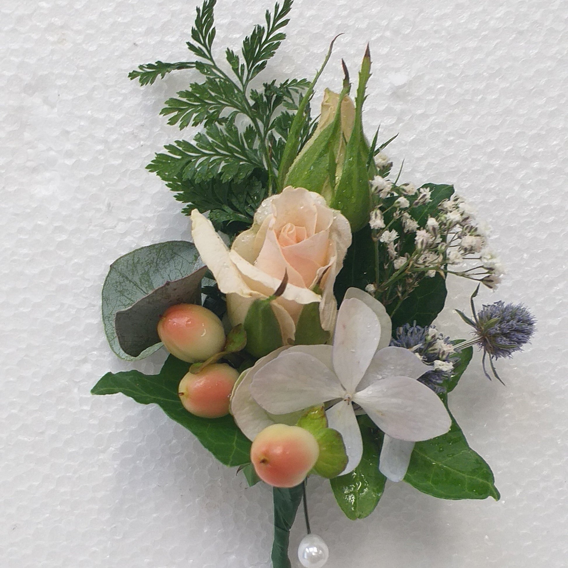 A small floral boutonniere with white orchid, pale peach rose, green foliage, and small orange berries, attached to a green stem and photographed against a white textured wall.