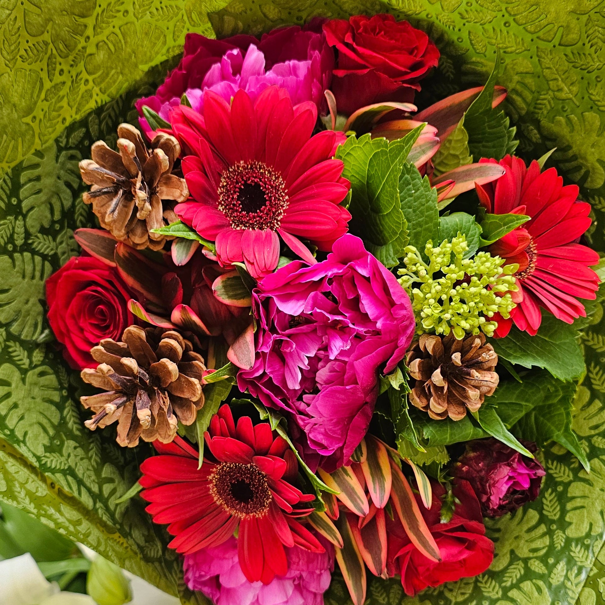 A vibrant flower bouquet wrapped in textured green paper, featuring bright pink and red gerbera daisies, red roses, magenta peonies, green buds, and natural brown pinecones.
