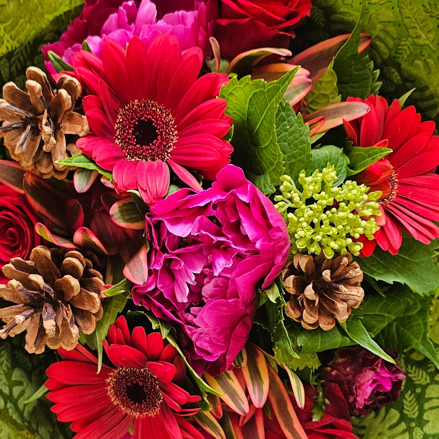 A vibrant flower bouquet wrapped in textured green paper, featuring bright pink and red gerbera daisies, red roses, magenta peonies, green buds, and natural brown pinecones.