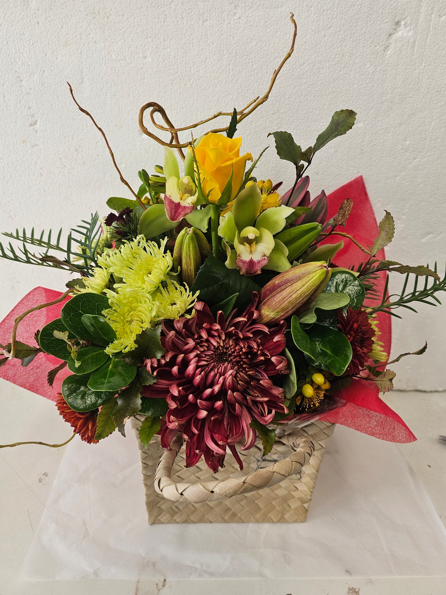 A mixed floral bouquet featuring a large burgundy chrysanthemum, yellow roses, green lilies, and light green feathery filler flowers, wrapped in red paper and tied with a fla woven basket, 