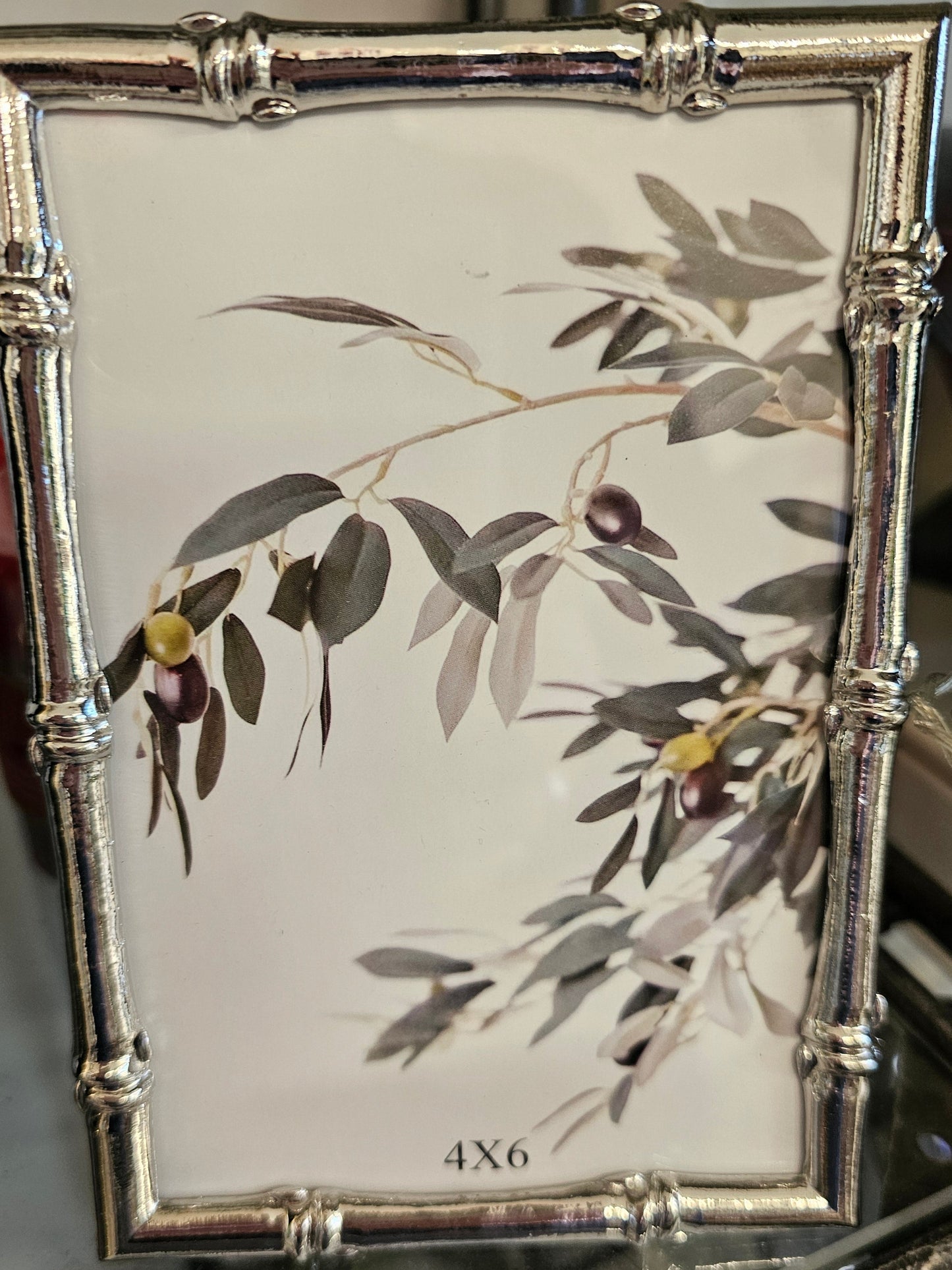 A rectangular picture frame with a silver-toned bamboo-style border and a printed olive branch design on a white background, shown hanging in a store setting.