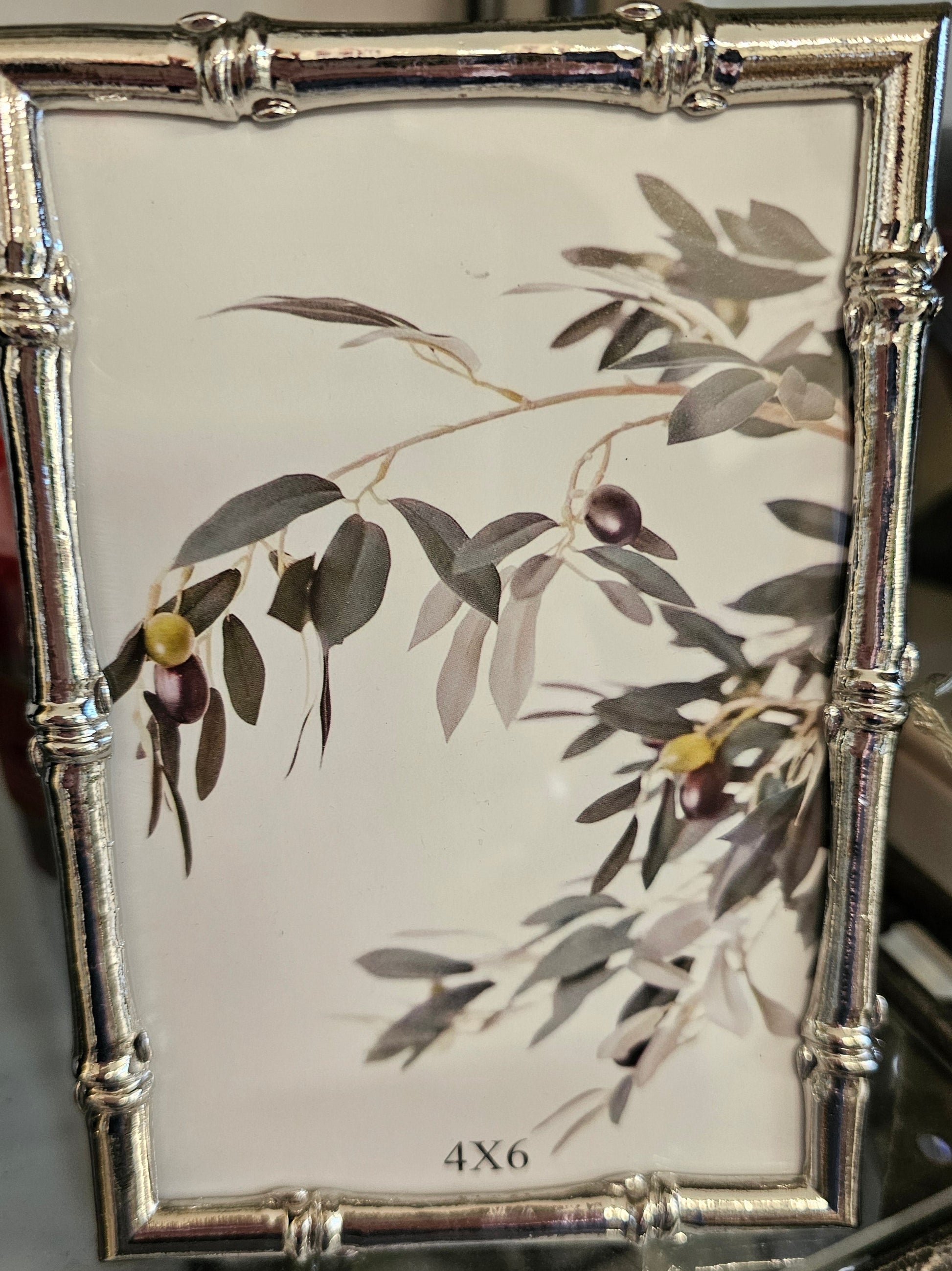A rectangular picture frame with a silver-toned bamboo-style border and a printed olive branch design on a white background, shown hanging in a store setting.