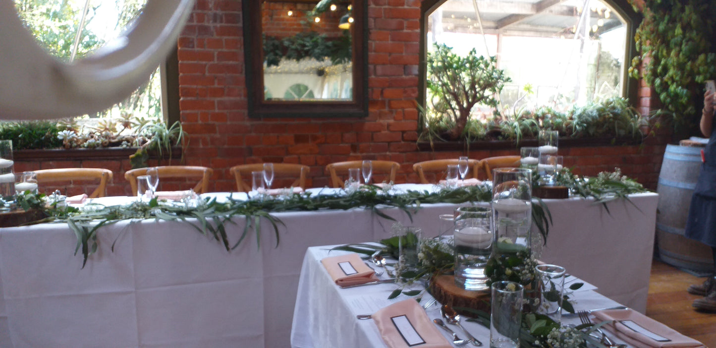 A rustic wedding reception table setup with white tablecloths, green leaf garlands, 